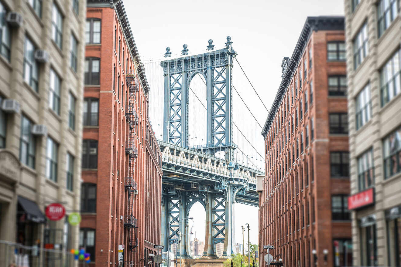 View of the Manhattan Bridge framed by tall red brick buildings on either side, with a distant city skyline visible beneath the bridge.